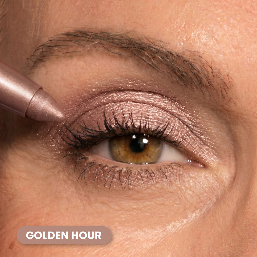 A close-up of an eye with shimmery golden-pink eyeshadow being applied with a makeup pencil. Eyebrow and skin texture are visible. The words GOLDEN HOUR are displayed in the corner.