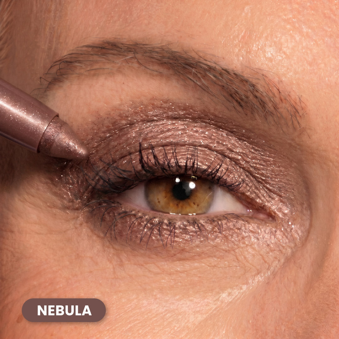 Close-up of a persons brown eye with metallic mauve eyeshadow applied to the eyelid. A matching makeup pencil is used along the upper lash line. The word NEBULA appears in a label at the bottom left.