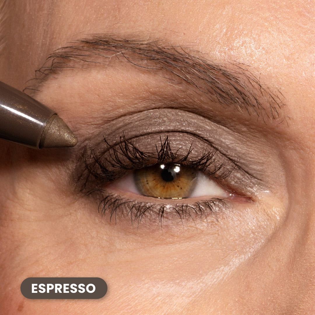 A close-up of a persons eye with light brown eyeshadow and brown eyeliner applied; a brown eyeliner pencil is held near the outer corner. The word Espresso appears in a label at the bottom left.