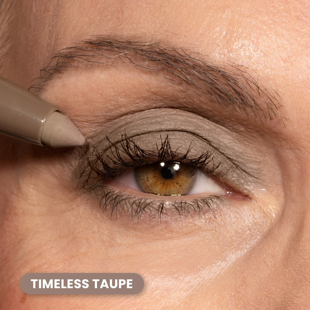 A close-up of an eye with taupe eyeshadow being applied to the upper eyelid using a makeup pencil. The product shade is labeled Timeless Taupe in the lower left corner.
