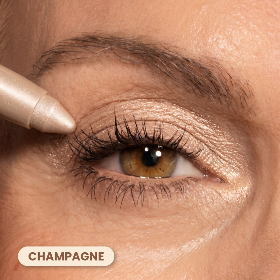 A close-up of an eye with champagne-colored eyeshadow applied to the eyelid. An eyeshadow pencil is being used on the upper lash line. The word CHAMPAGNE appears at the bottom left.