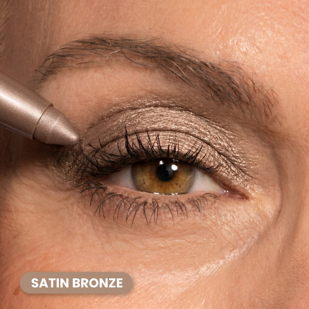 A close-up of a persons eye with bronze eyeshadow applied to the eyelid. An eyeshadow pencil is touching the outer corner. The text SATIN BRONZE appears at the bottom left of the image.