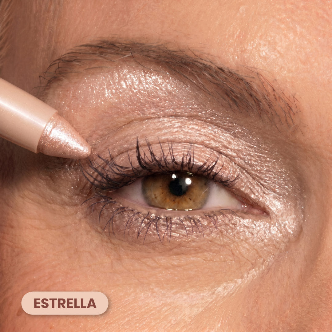 A close-up of a persons eye with shimmering, light pink eyeshadow. A matching creamy eyeshadow pencil is being applied to the upper eyelid. The label ESTRELLA appears in the bottom left corner.
