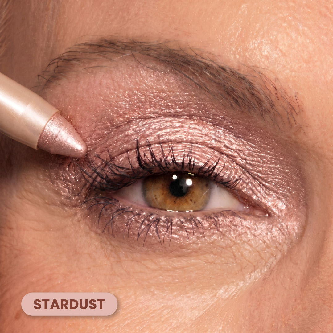 A close-up of an eye with shimmery pink eyeshadow and eyeliner being applied with a makeup pencil. The eyeshadow shade is labeled STARDUST in a rounded beige box at the bottom left.