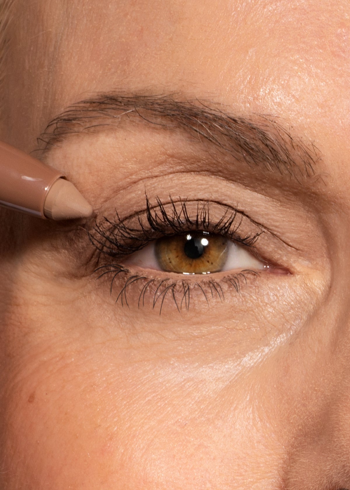 A close-up of a persons eye with brown iris, defined lashes, and a beige eyeliner pencil being applied to the upper eyelid.
