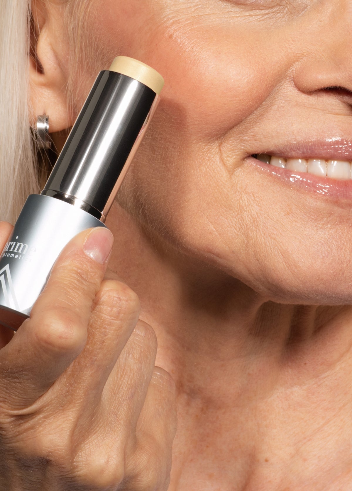 Close-up of an older woman smiling while holding a tube of stick foundation or concealer to her cheek. Her skin appears smooth and healthy, and she has light makeup on.