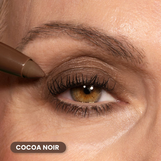 A brown eye with long lashes is shown as a person applies dark brown eyeliner to the upper eyelid. The shade is labeled Cocoa Noir in white text on a brown background at the bottom left.