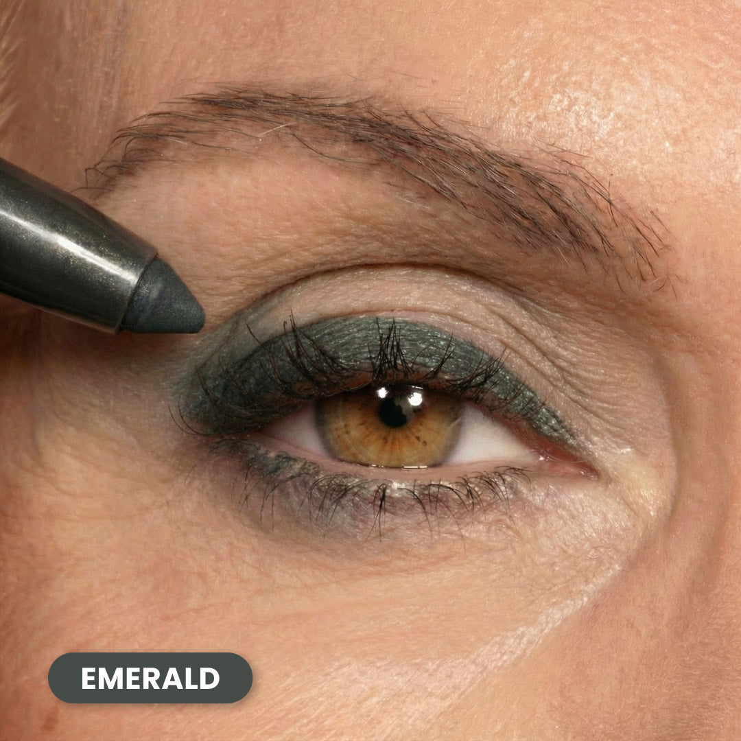 A close-up of a persons eye with green emerald eyeliner applied to the upper and lower eyelids. An eyeliner pencil is positioned near the outer corner of the eye. The word EMERALD appears in a label at the bottom left.