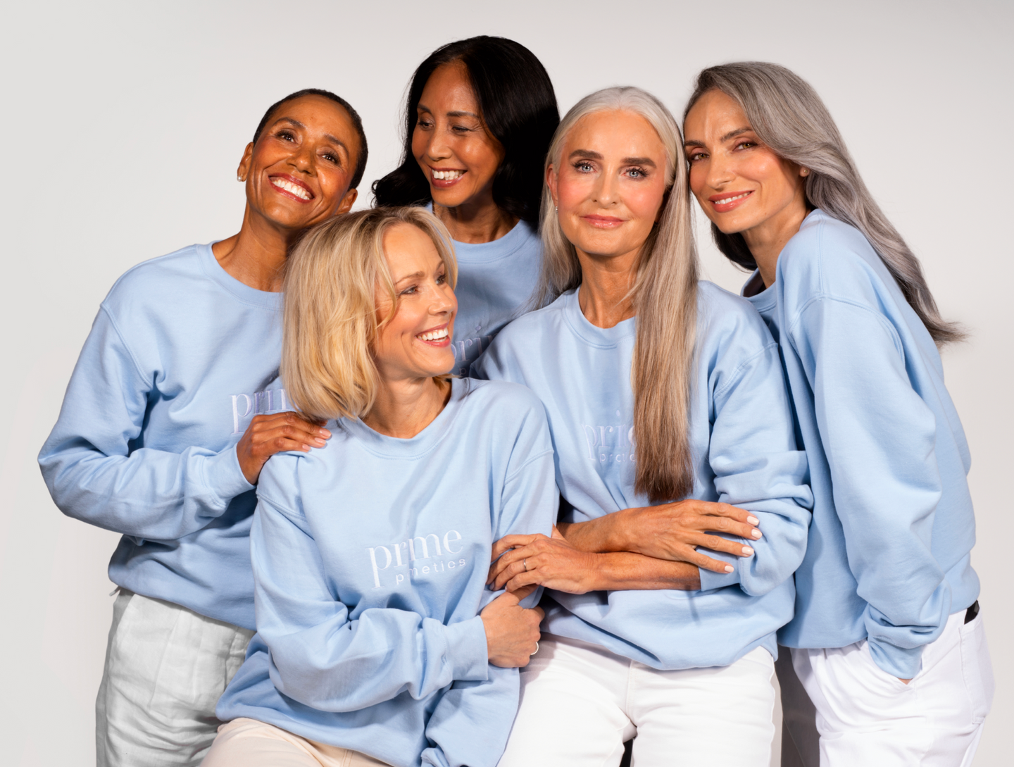 Five women with gray and blonde hair, wearing matching light blue sweatshirts and white pants, stand closely together and smile warmly against a plain background.