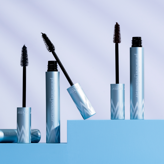 Three blue tubes of volumizing mascara stand on staggered blue platforms, each with its black applicator wand exposed. The background is light with soft diagonal shadows.