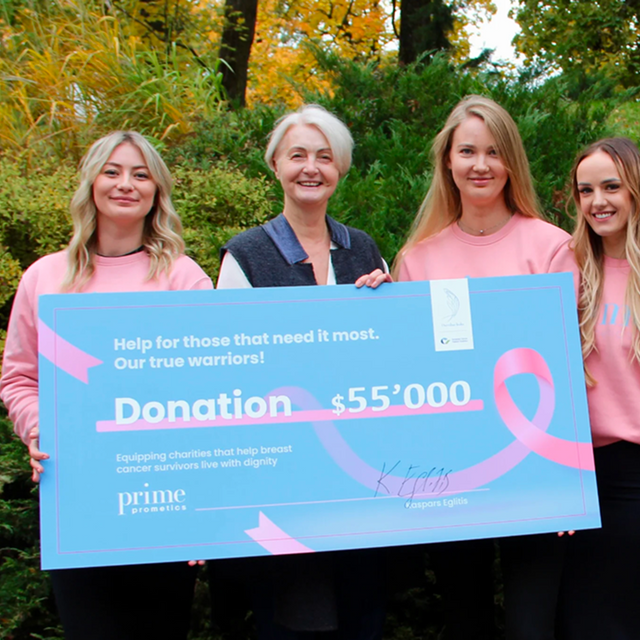 Four women, smiling, stand outdoors holding a large check for $55,000, donated to support breast cancer survivors. The check is blue with pink ribbons, and the women wear matching pink sweatshirts. Lush greenery is in the background.