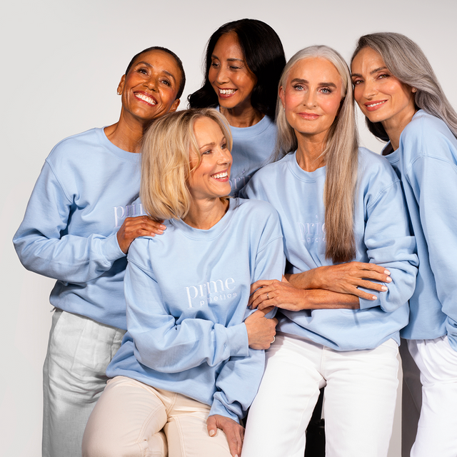 Five smiling women with diverse hair colors and styles, all wearing light blue sweatshirts and light-colored pants, pose closely together against a plain white background.