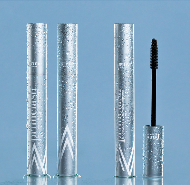 Three silver mascara tubes, one with the wand exposed, are standing upright on a reflective blue surface. The tubes are covered in water droplets, giving a fresh, dewy appearance.