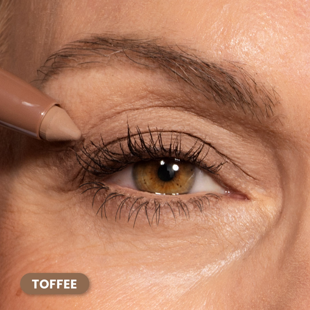 A close-up of a persons eye as they apply a toffee-colored makeup pencil to their upper eyelid. The eye has brown and green tones, and the eyebrow and eyelashes are clearly visible. A label reads TOFFEE.