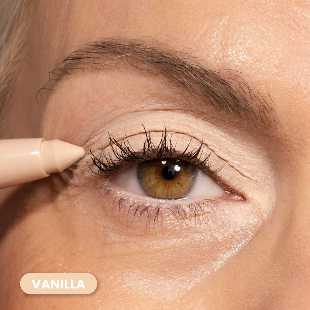 A close-up of a persons eye with light skin, applying a beige eyeliner pencil along the upper lash line. The shade is labeled Vanilla in a rounded box at the bottom left.